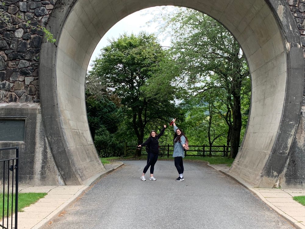 Two people stood posing with their arms up and out under a stone arch.