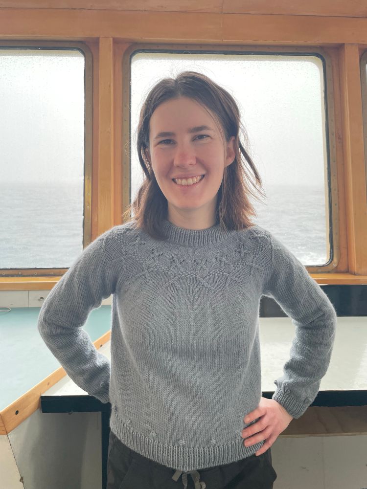 Photo of a white woman with brown hair wearing a hand-knit sweater. The sweater is light blue, has a design intended to look like Chaetoceros diatoms around the yoke, and bobbles around the waistline and wrists