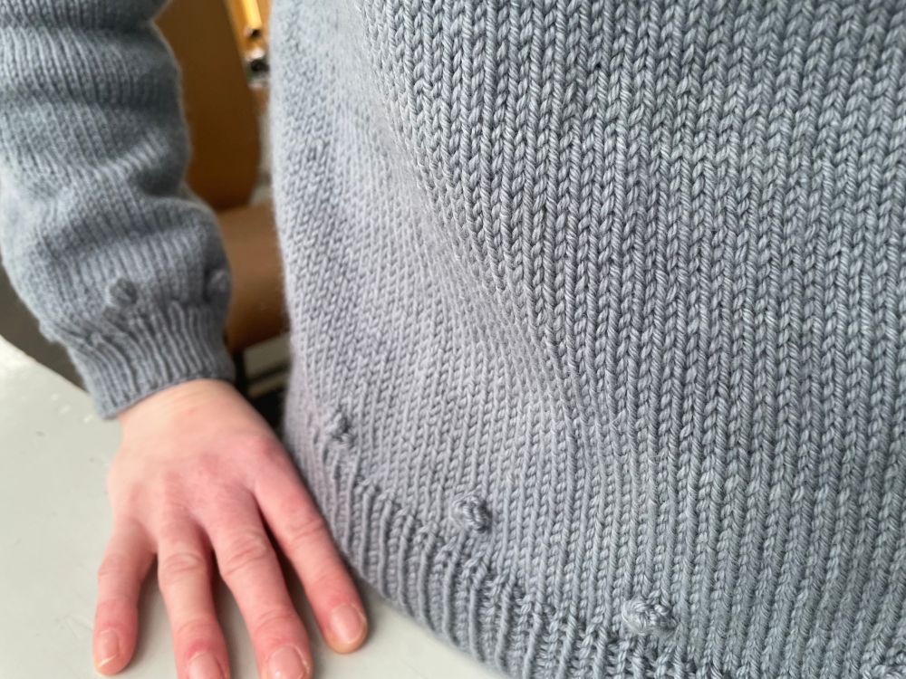Photo of a hand-knit light blue sweater, zoomed in on the bottom hem. Small bobbles are distributed around the wrist and waistband, representative of Chaetoceros resting spores