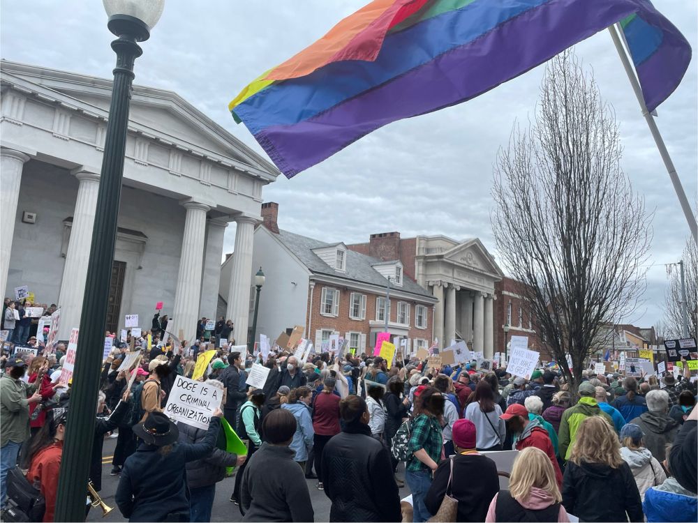 Photo of a large crowd outside of a city hall building. Many people hold up signs in protest and a rainbow flag waves in the top right corner