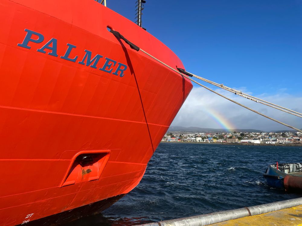 Photo of the Nathaniel B Palmer research vessel in port in Punta Arenas. The ship’s orange bow with PALMER written in blue is on the left side, and the sea, the town, and a rainbow are on the right side