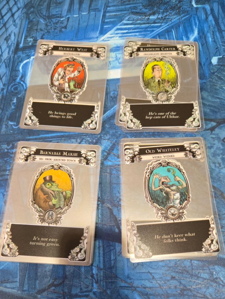 Top cards in each stack, starting upper left, going clockwise: Herbert West Reanimator "He brings good things to life", Randolph Carter Dreamland Believer "He's one of the hep cats of Ulthar", Old Whateley Weird Wizard"He don't keer what people think", and Barnabas Marsh Big Frog Around Town. "It's not easy turning green"