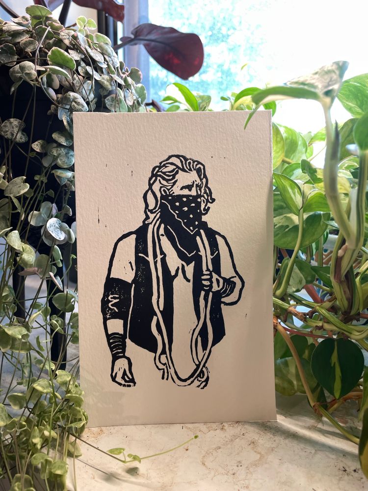 a black block print of hangman adam page on white cardstock, propped up against house plants. hangman is wearing a bandanna covering his face, a black vest, one elbow pad, and has rope around his neck