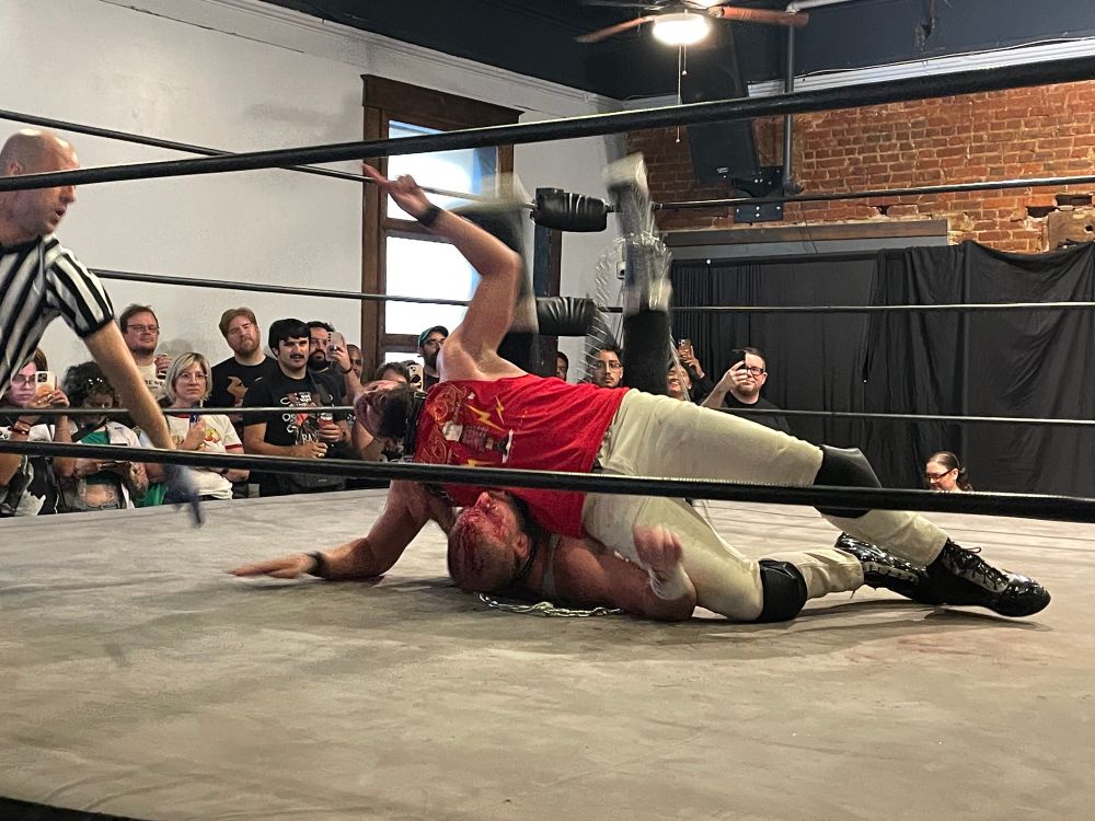 drew gulak connected by a dog collar and chain to brian johnson 'the mecca'. the mecca is pinning a cut open gulak with his whole body