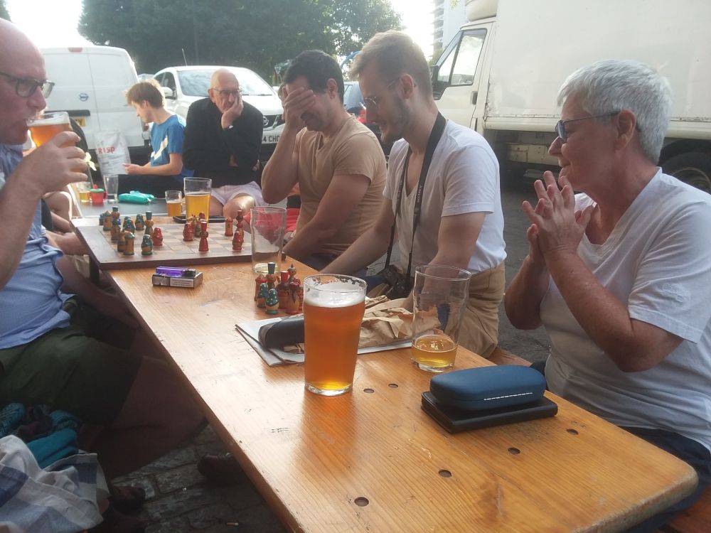 A group of people sitting at tables in the sunshine, drinking beer, chatting and playing chess. The person sat at the chess board has his hand over his eyes after making a mistake, while his opponent sips his beer. In the background people are deep in conversation, with one resting his chin on his hand.