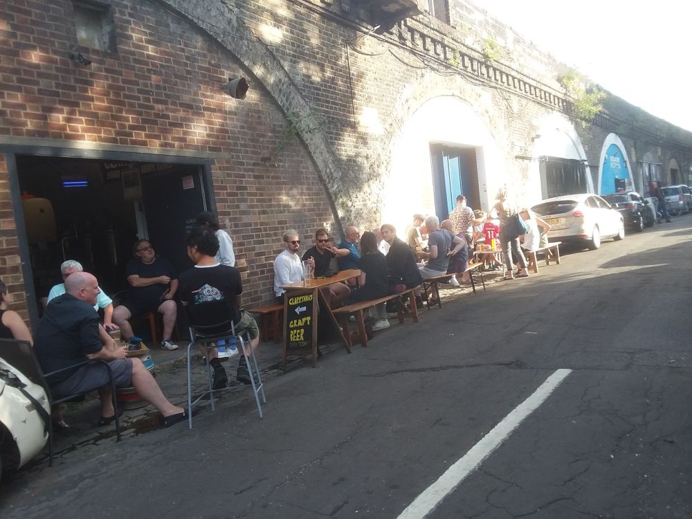 People standing and sitting at tables and chairs outside a railway arch in sunshine and dappled shade, with a curve of arches and parked cars  into the distance. A sign says Clarkshaws Craft Beer with an arrow pointing towards the doorway. 