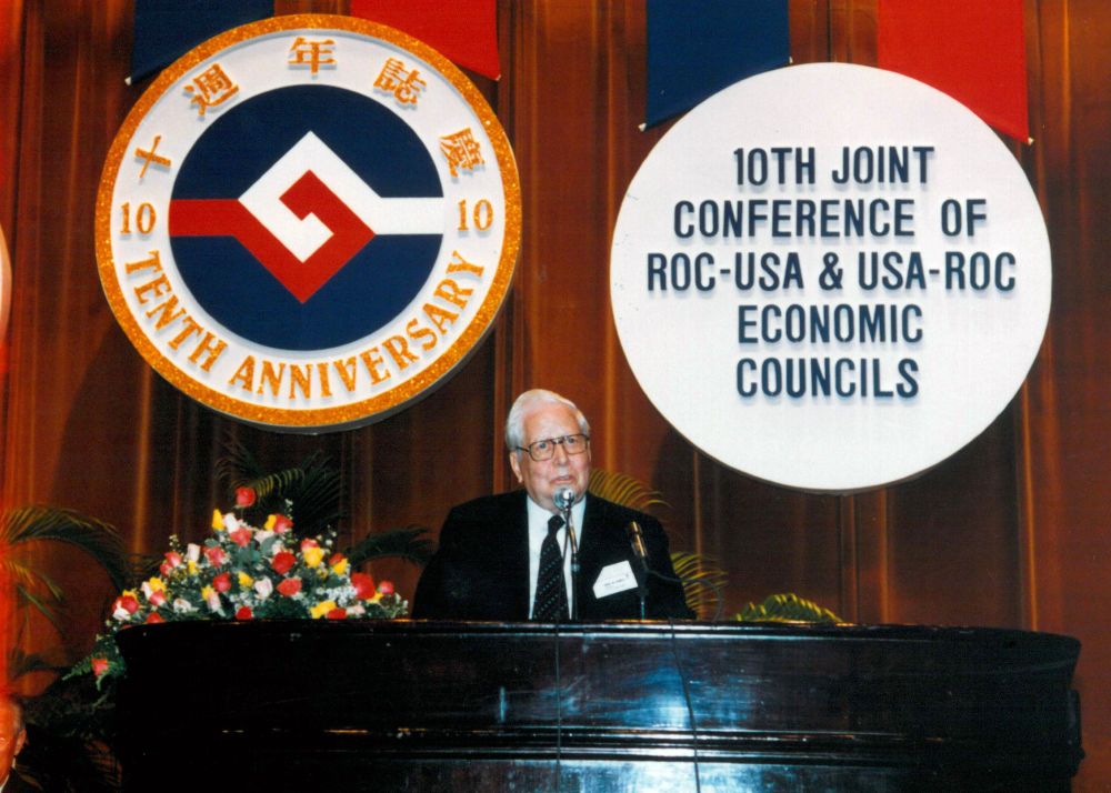 Chairman David M. Kennedy at the 10th joint business conference of the USA-ROC and ROC-USA Economic Councils. In the back on red pipe and drape are the joint anniversary logo on the left, and text commemorating the event on the right, with Chairman Kennedy on a table next to a flower arrangement.