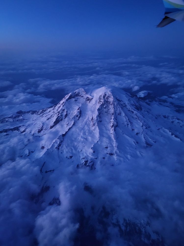 Aerial photo of Mt. Rainier covered in snow and surrounded by clouds. Twilight casts a glow over the area.