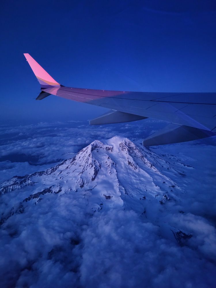 Aerial photo of snowy Mt. Rainier under the wing of a plane. The sun is setting, casting a twilight glow over it and the clouds surrounding it.