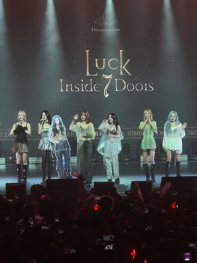 Dreamcatcher no Brasil, em 15/09/24. Seis integrantes aparecem interagindo com o público à frente, com exceção de Dami, que está sendo representada por uma imagem de papelão em tamanho real e segurada por JiU. Ao fundo o nome da turnê, Luck Inside 7 Doors.