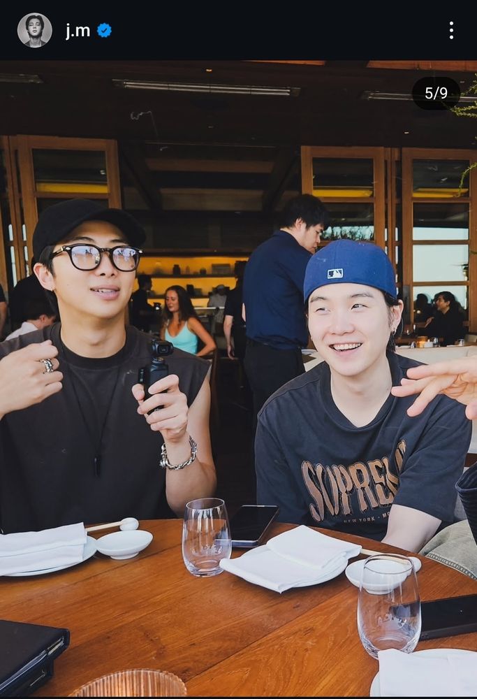 Foto de Namjoon e Yoongi, sentados à mesa. Namjoon segura uma câmera  e Yoongi está sorrindo. 
