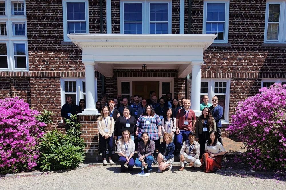 VCID Conference attendees posing in front of a brick building with large blooming pink shrubs on a sunny day at The Jackson Laboratory in Bar Harbor, Maine.