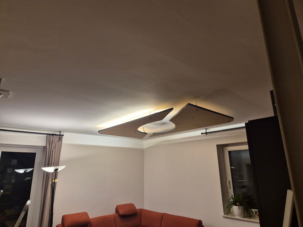 Selbstgebaute Deckenlampe im Wohnzimmer aus massiven Holzbrettern die passives Licht in Warmweiß ausstrahlt - zumindest auf der linken Hälfte. 
Die Lampe besteht aus einer rechteckigen Holzplatte die diagonal zersägt wurde, mit einem ovalen Loch in der Mitte. 
Sie hängt etwa 10cm unter der Decke. 