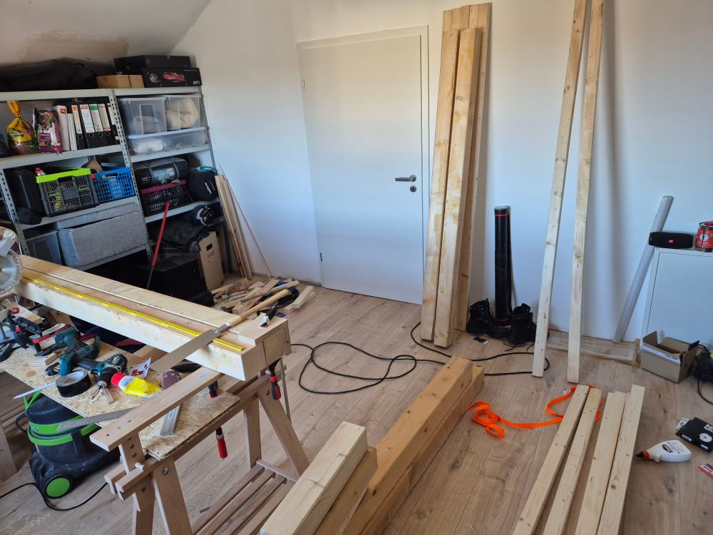 Werkraum in dem mehrere große Holzbalken auf dem Boden liegen. einigere kleinere lehnen an der Wand, 2 weitere zusammen mit Zollstock und Japansäge auf Böcken. Im Hintergrund ein Lagerregal mit Kisten und Ordnern.