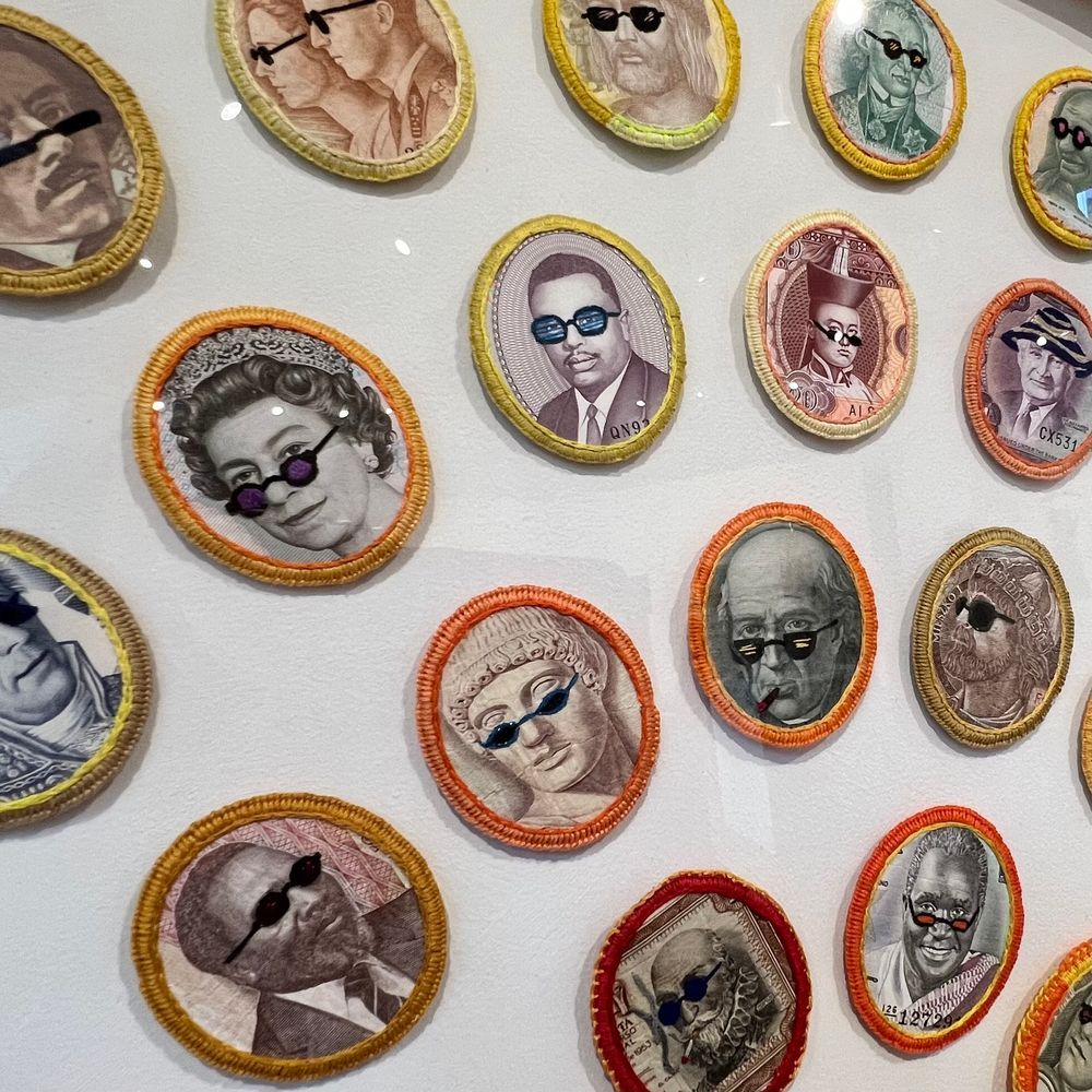 A close up of some of the embroidered international currency portraits in artist Stacey Lee Webber's colorful piece. Historical figures don stitched fun glasses and hats.