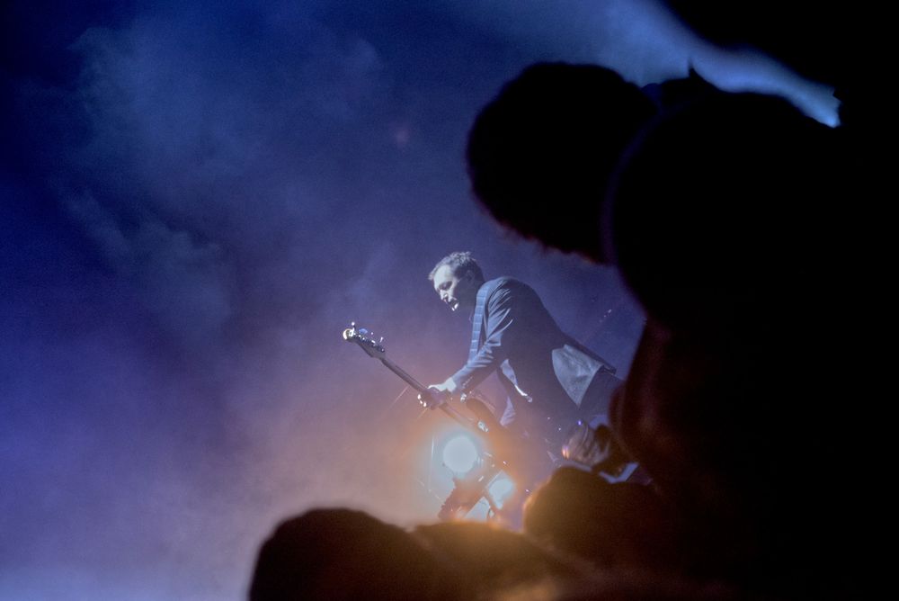 A angled photo of a guitarist from Interpol playing with a bright light behind him and a blurry crowd in the foreground
