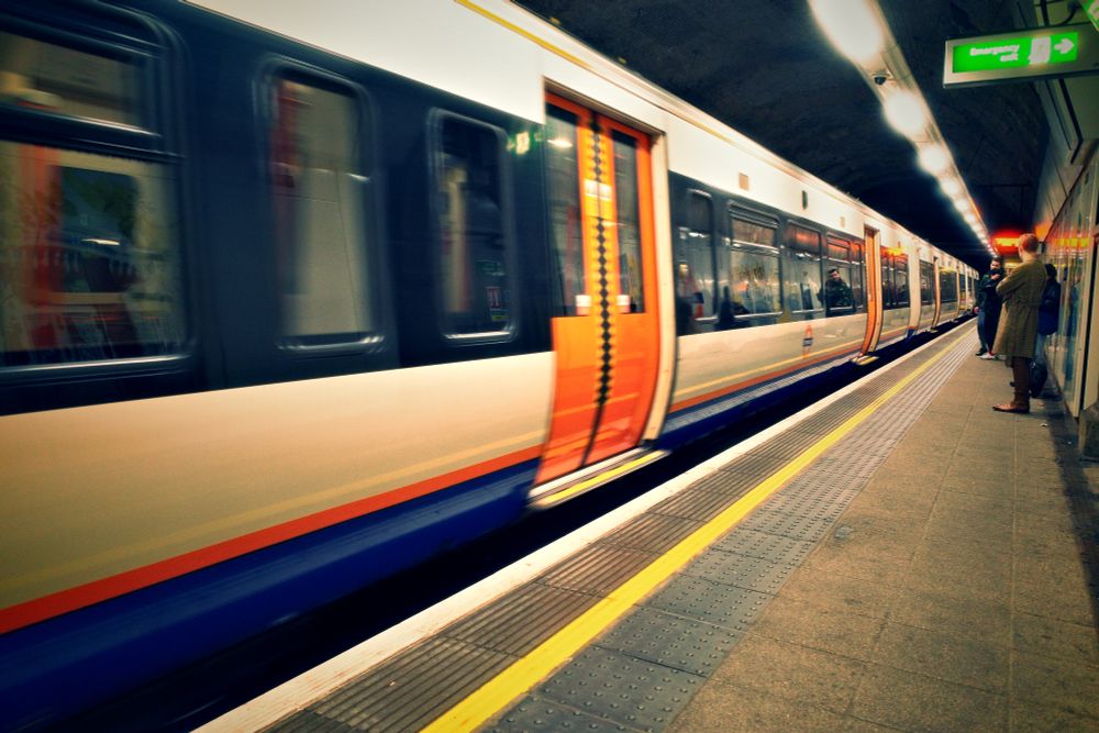 A train passing a London Underground paltform. To the right of frame there are some people waiting on their train.
