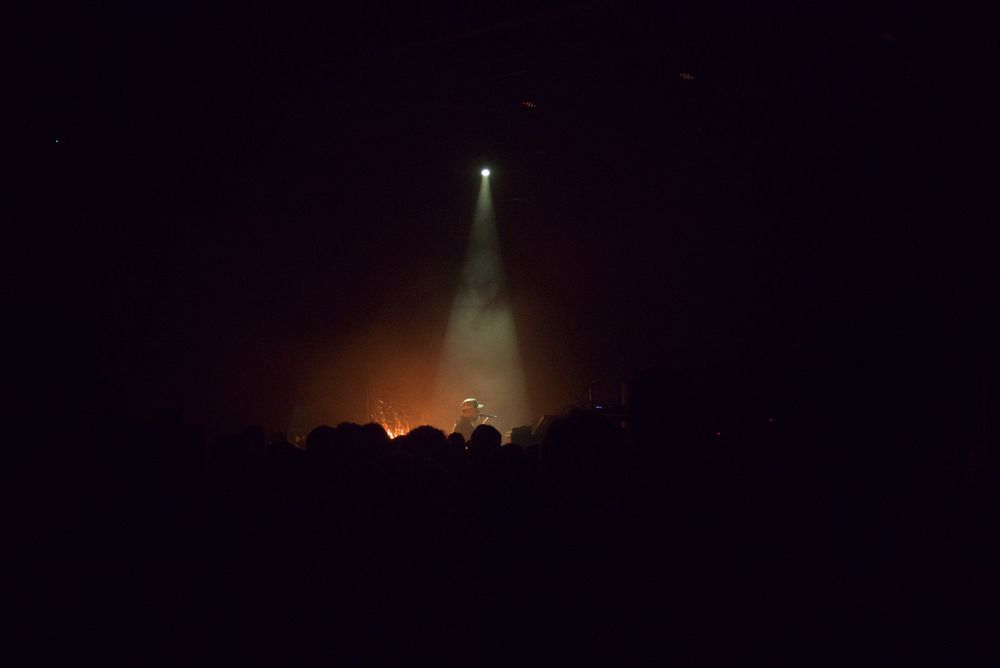 A photo of John Grant playing live in Vicar Street singing into a microphone illuminated by a spot light with the crowd visible in the foreground