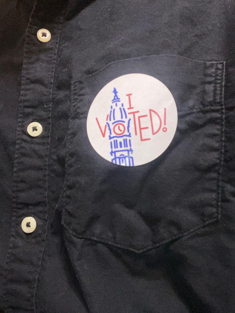 A close up of one side of a black button down shirt, on its front pocket is a sticker with the letters of “I VOTED” intercut by an image of the clock tower of the city hall of Philadelphia, where the O in voted is placed in the spot where  the clock would be and contains clock hands.
