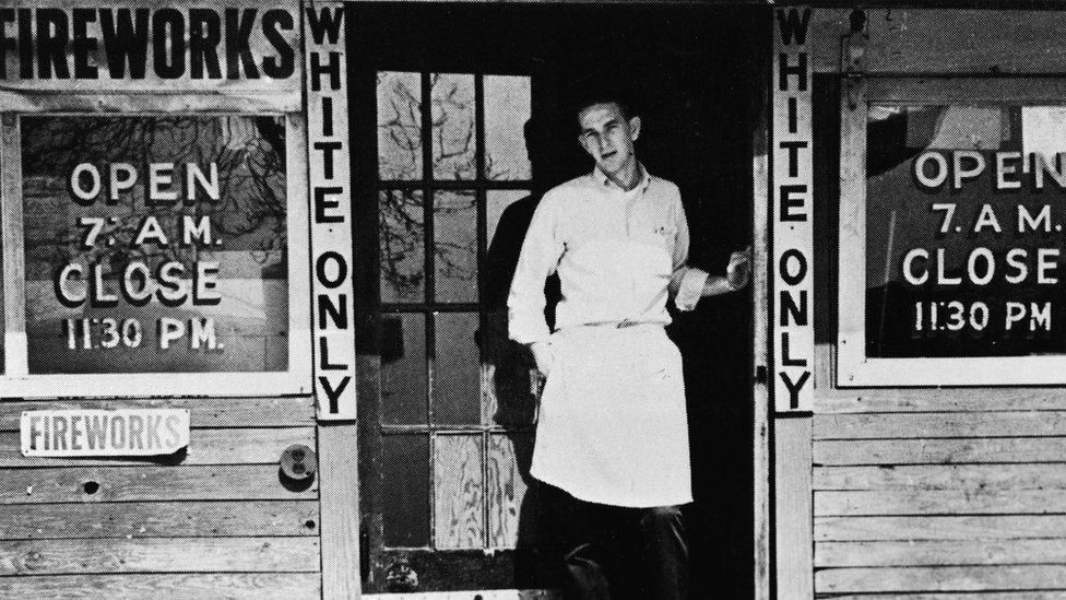 A proprietor of a fireworks shop (?) standing in the door of his establishment, on either side of which there are signs saying "WHITE ONLY"