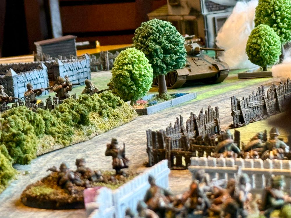 Photograph of a tabletop wargame set in World War II. An enemy tank appears around a tree up the road while friendly infantry huddles behind wooden fences