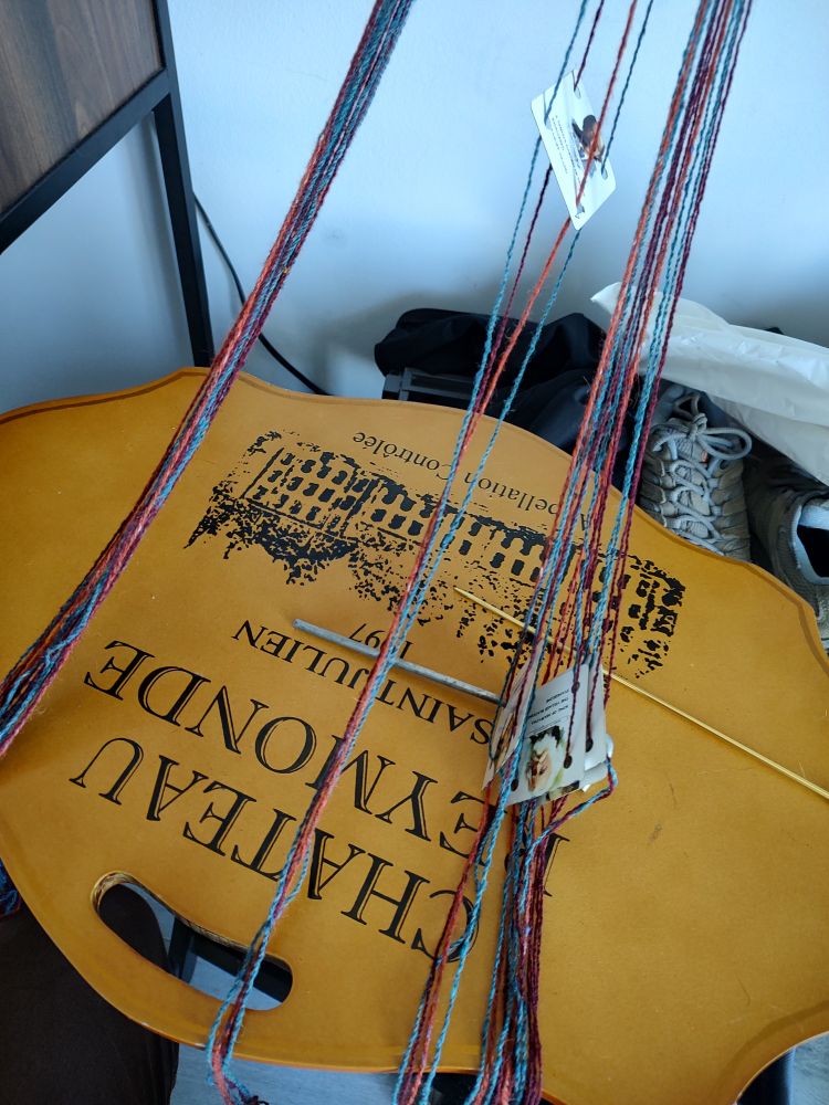 a half-organized tangle of blue, orange, and maroon yarn partially strung onto weaving tablets cut from a deck of "famous authors" playing cards. the tablets are held in place by a steel tent stake. The entire assembly is coming together into something almost, but not quite, entirely unlike a backstrap loom