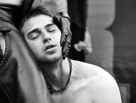 A naked slave kneels next to its owner, resting its head on its owner's leather trousers, its eyes are closed and it looks content