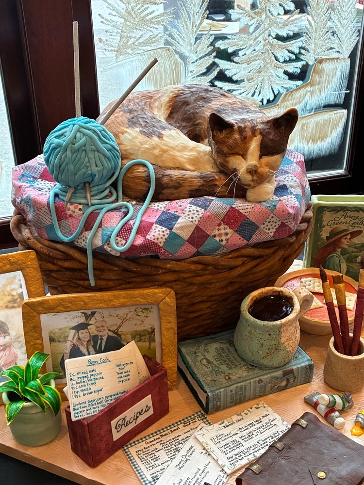 A gingerbread cat napping in a knitting basket surrounded by pictures, recipe cards, and various desktop items. 