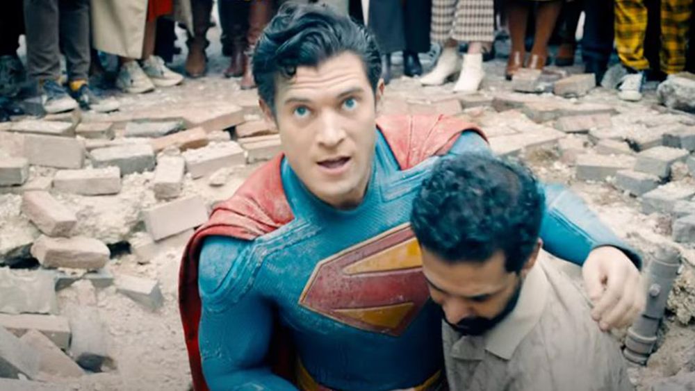 Superman is helped out of a crater by an onlooker. Superman looks to the sky.