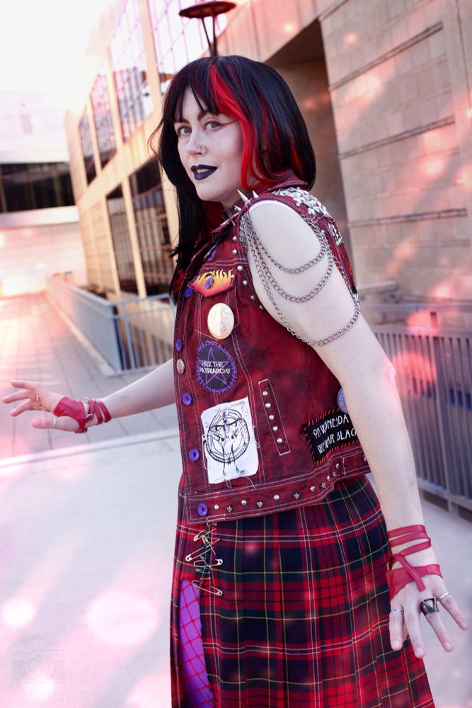 Image of a cosplayer wearing a costume that is a punk variant of Sailor Mars, posing turned towards the camera outside in late afternoon