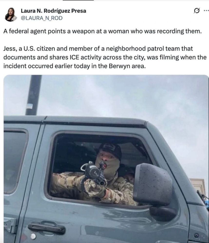 Screenshot of a tweet with a photo below. Visible text:
“Laura N. Rodríguez Presa
@LAURA_N_ROD

A federal agent points a weapon at a woman who was recording them.

Jess, a U.S. citizen and member of a neighborhood patrol team that documents and shares ICE activity across the city, was filming when the incident occurred earlier today in the Berwyn area.”

Photo description: In the attached photo, a masked person in camouflage sits in the driver’s seat of a gray SUV, pointing a firearm out the open window toward the camera under a gray sky.
