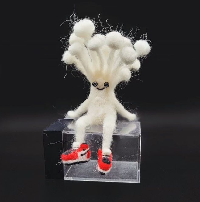 A white mushroom guy with multiple little sprouts growing from the top of his head, sitting on an empty dice box. His feet are hanging over the side. He's wearing little red jordans.