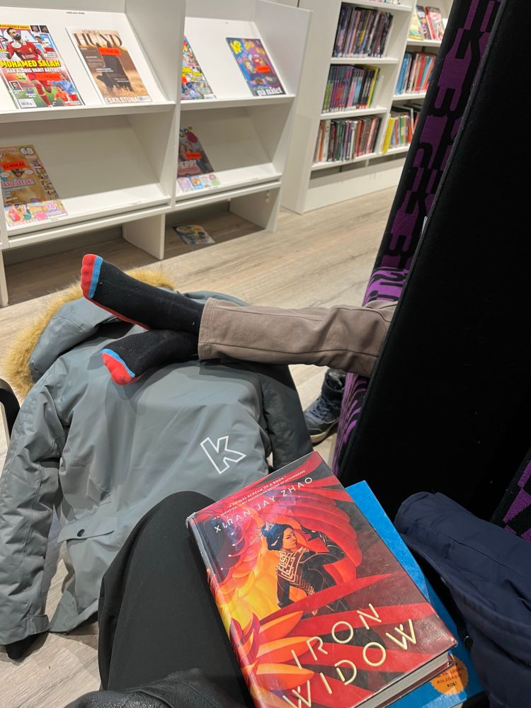 Holding the book Iron Widow by Xiran Jay Zhao. My 10 year old’s legs are sticking out from a cubicle chair and resting on top of a stool covered by his jacket, as he sits and reads. Bookshelves and magazine racks are in the background.