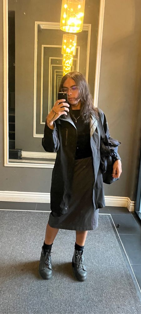 apartment lobby selfie of me wearing a black trench with a black top and gray midi skirt and boots