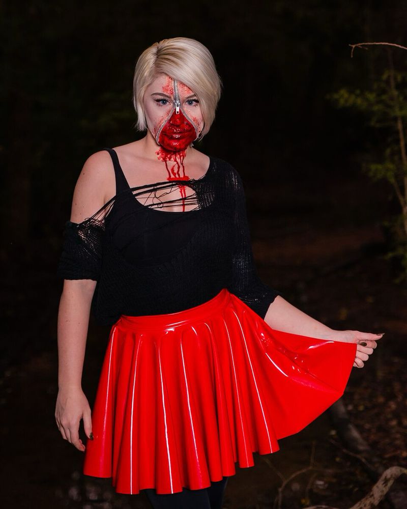 A closer shot ofwhite woman with short blonde hair staring, wearing a shredded black jumper and bright red pvc skirt, with a zipper on her face revealing bloody gore underneath 