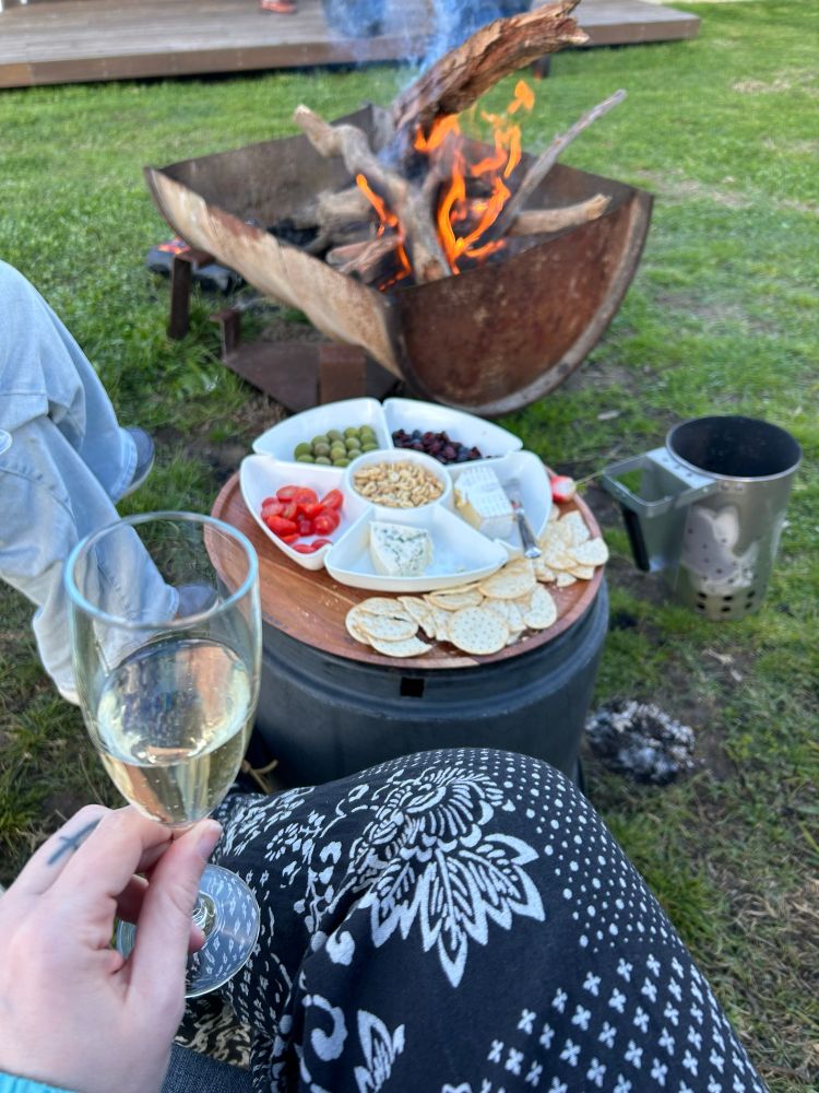 A champagne glass is held closer to frame while in the background a campfire and charcuterie can be seen 