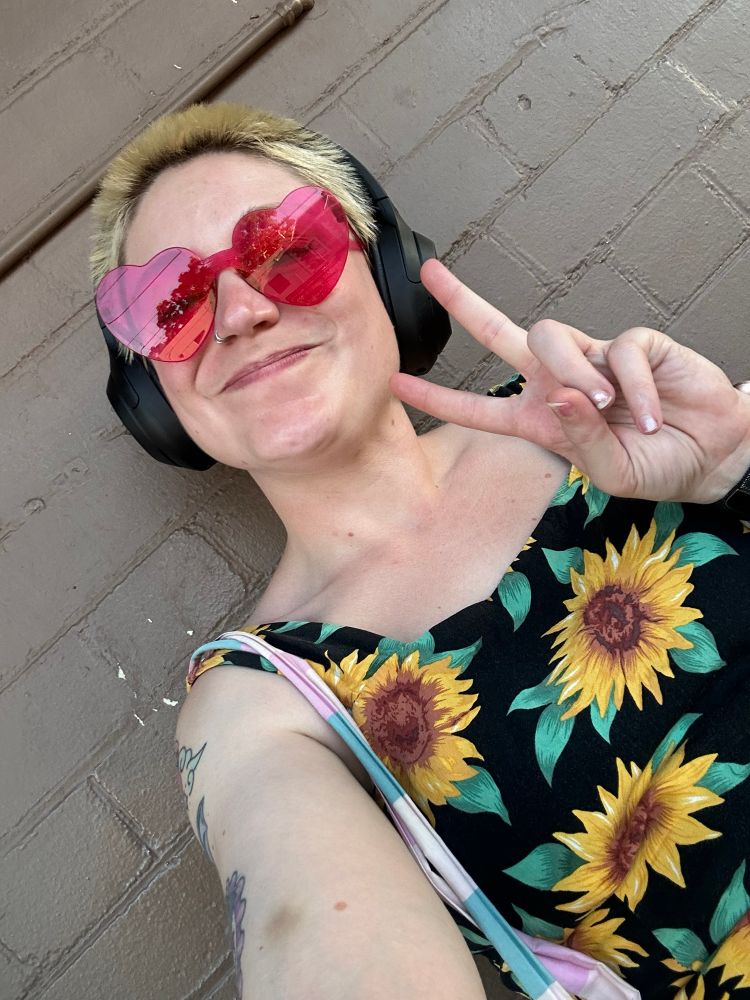 A white gal (OP) poses with her hand held in a peace sign. Smiling and wearing heart shaped pink sunglasses, she is wearing a sunflower jumpsuit