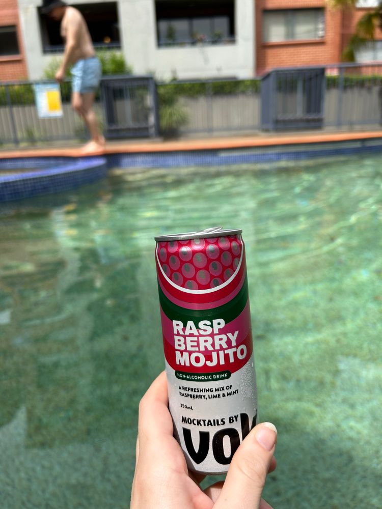 A raspberry mojito mocktail can is held in the foreground of the photo while the background shows a pool and sunshine