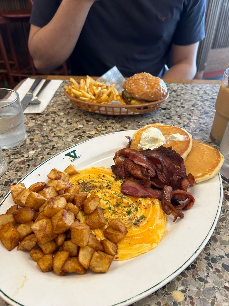 A delicious breakfast sits in front with pancakes, eggs, home fries, and bacon, along with two iced coffees. Further back on the table is a cheeseburger and fries. Everything was delicious. 