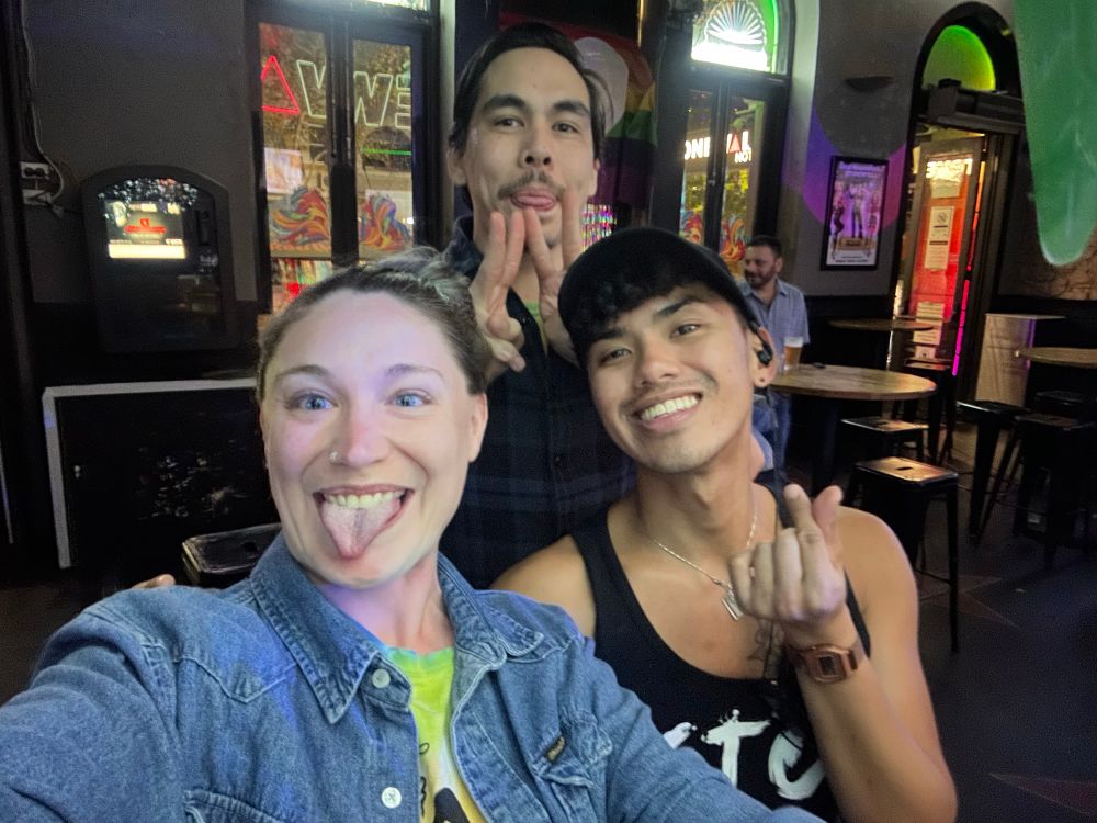 Three folks grin at the camera. Op is a white woman with purple hair sticking out her tongue on the left of the frame. Her partner is behind her making a silly face, and Jeff the best bartender is on the right and is so slay