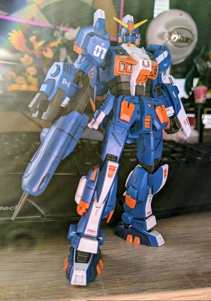 A blue, orange, and white mecha (the Gundiver) poses heroically, on a desk, since it is a model kit.