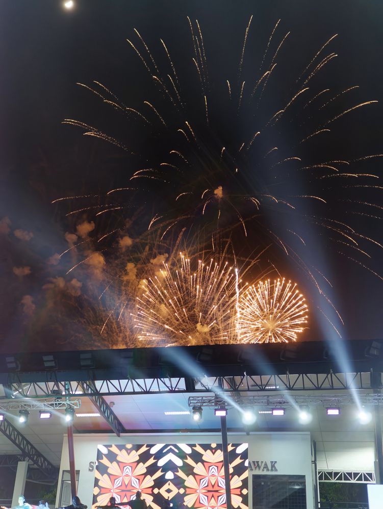 multiple fireworks lit the night sky with moon, behind a hokey stadium grandstand 