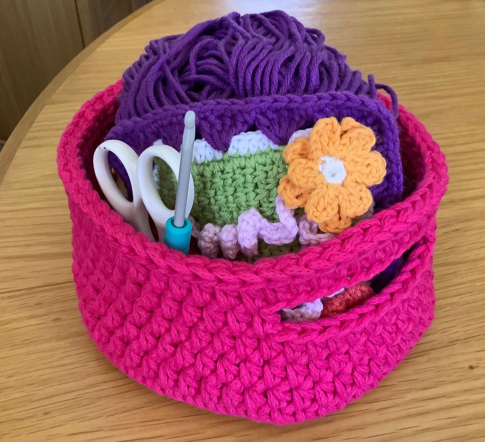 My Fuscia-coloured chunky, round crochet work basket with integral handles, containing hook, scissors, purple yarn and an arty 3D flower granny square that I made during the pandemic.