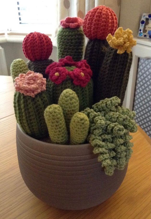 Grey coloured curvy flower pot filled with various types of crochet cacti & succulents. Some tall, dark green ‘ribbed’ types, with little yellow and pink flowers on the top and some lighter green, smooth and round, others spiral shaped, dangling over the side of the pot.