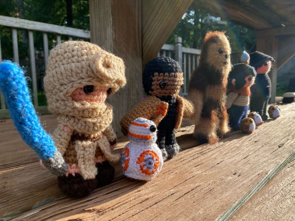 Crocheted figures of Rey, BB-8, Finn, as Chewbacca , Rose Tico and her Porg friends and Ben Solo. They are standing in a line on a wooden porch, looking to the right. The sun is shining on them. 