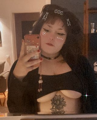 Uma foto minha, acho que de 2022, na frente de um espelho com um body de decote invertido e blusa de manga longa de cor preto. Cabelo médio escuro. Tatuagem de girassol na barriga debaixo dos seios.