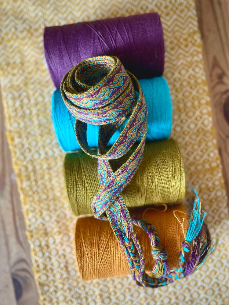 a woven band with alternating dragon heads is spread out in a spiral atop the 4 cones of cottolin in colors ockre, green, sky blue and purple that were used to weave the band. a woollen tablerunner  with a yellow rosepath serves as artsyfartsy bokeh. 