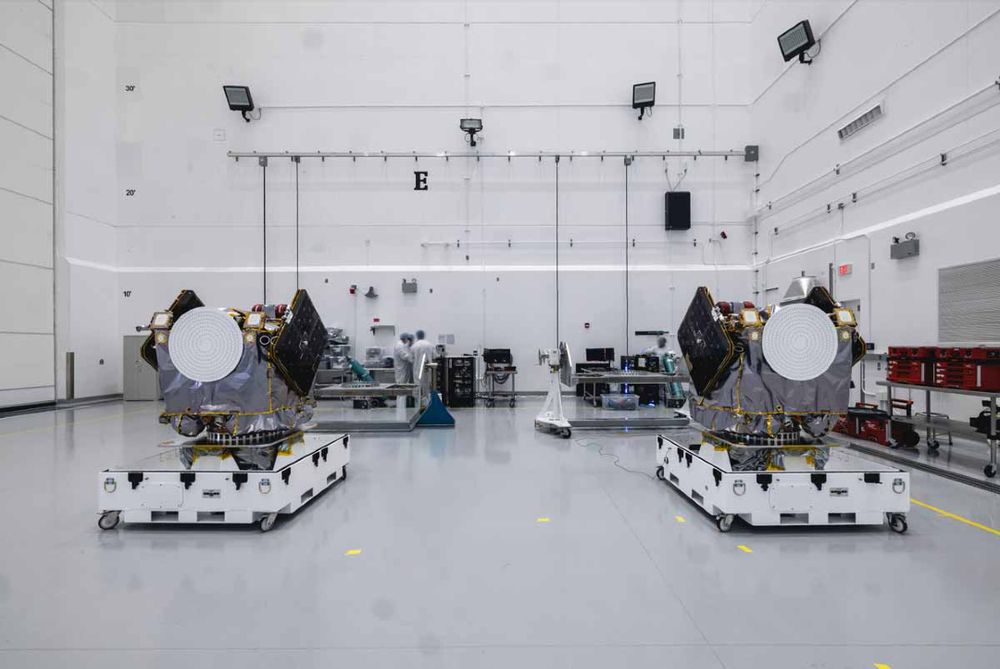 Deux satellites identiques sur des plateformes roulantes dans une salle blanche, entourés d'équipements techniques et de personnel en combinaison.