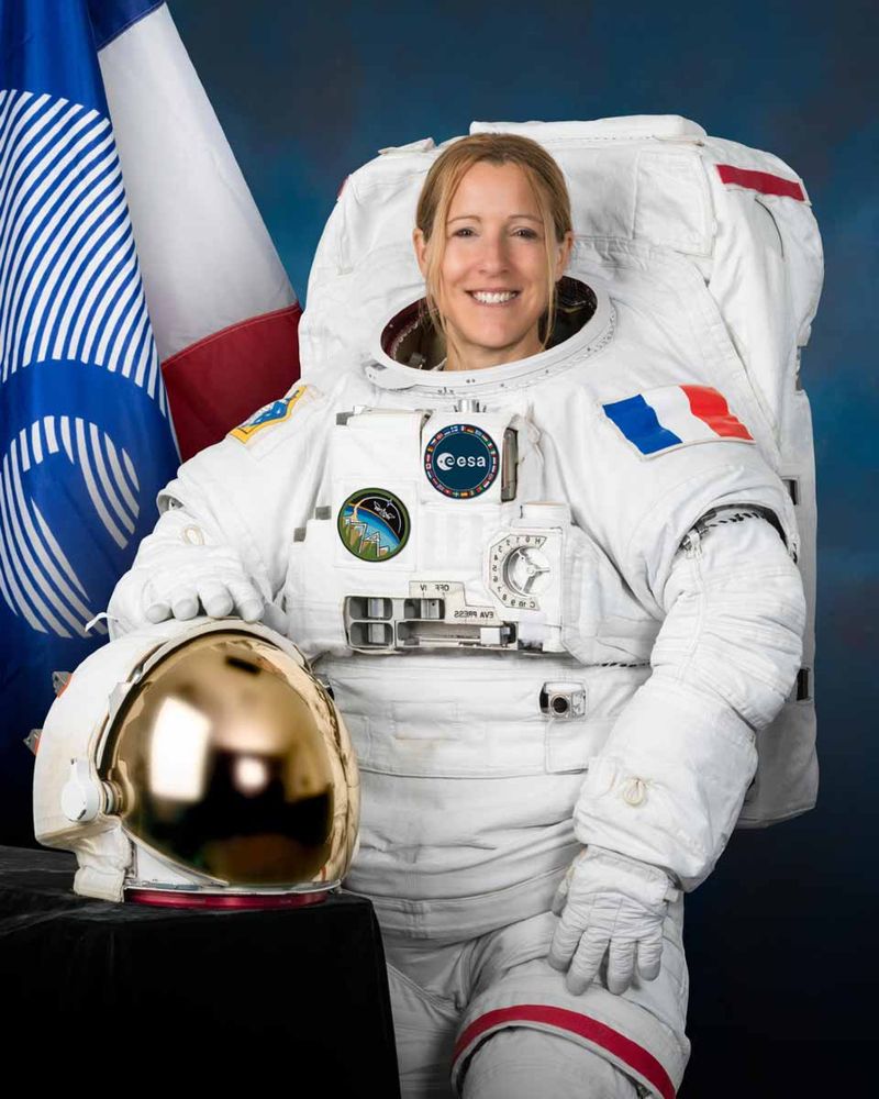 Sophie Adenot souriante en combinaison spatiale blanche, tenant un casque doré, avec drapeaux de la France et de l'ESA en arrière-plan.