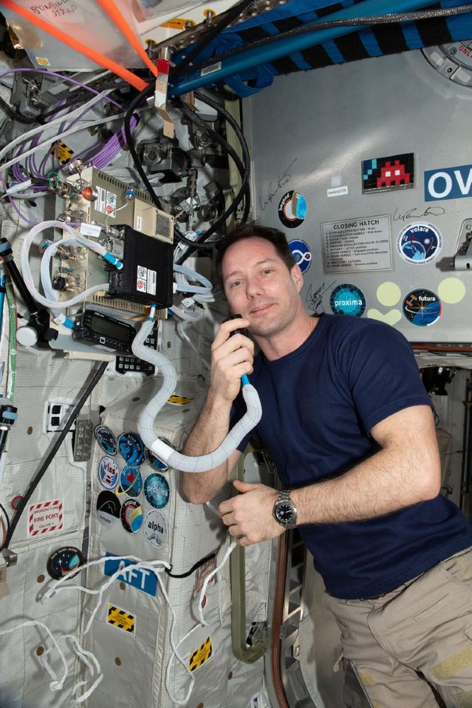 Thomas Pesquet, astronaute français, utilise un appareil de communication à bord de la Station spatiale internationale, entouré d'équipements et d'autocollants de mission.